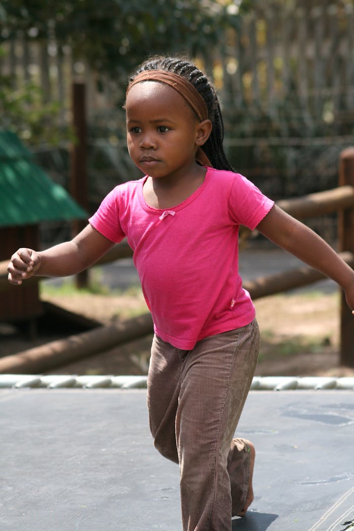 about-01 A young black girl in a pink shirt is running on a trampoline in a sunny outdoor setting.